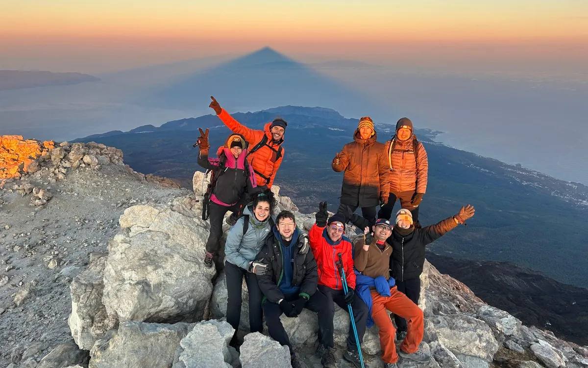 Hikers in down jackets on pale rocks at first light, the pointed shadow of the mountain behind them
