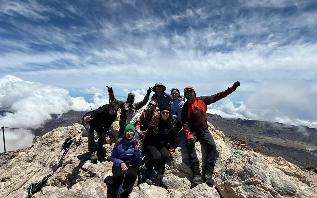 Eight hikers in hats, gloves and warm jackets on light rocks at the top, arms raised