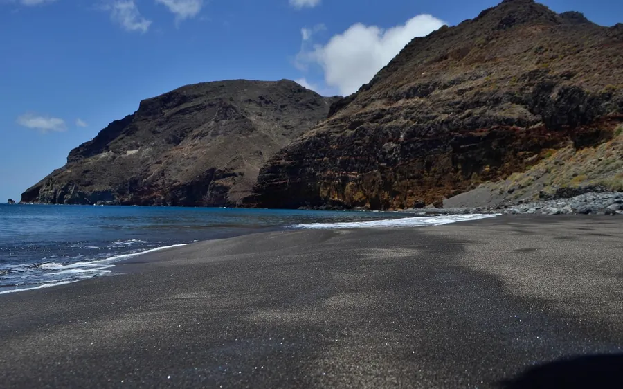 Ein Strand mit schwarzem Sand unter steilen braunen Landzungen und blauem Meer