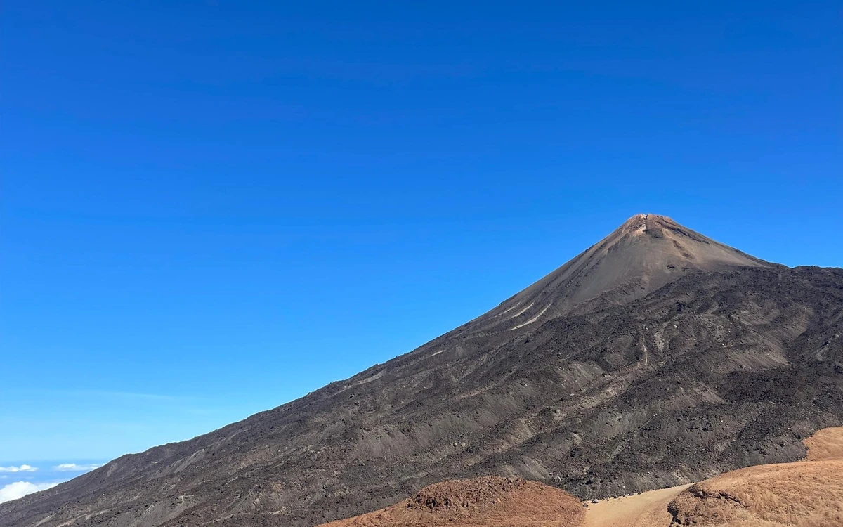 The grey cone of Teide against a deep blue sky above a slope of dark old lava