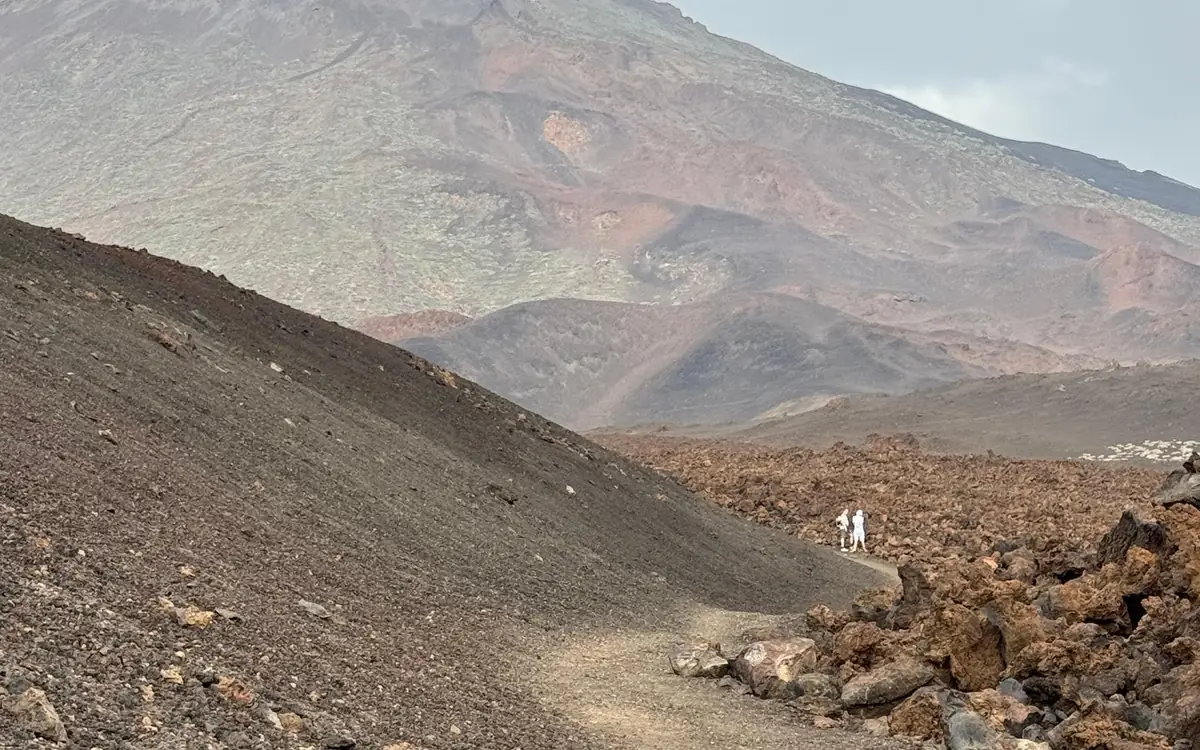 A sandy path between a dark cinder slope and a field of rust-brown lava blocks, two walkers far ahead